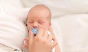 Śpiący noworodek z niebieskim smoczkiem w buzi. Rodzic delikatnie przytrzymuje smoczek, martwiąc się o potencjalny krzywy zgryz od smoczka.