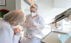 Dentystka w masce i fartuchu wykonuje zabieg kiretażu u pacjentki. W tle widać sprzęt stomatologiczny.