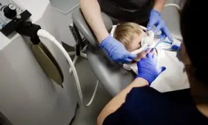 Chłopiec w masce do znieczulenia ogólnego u dentysty. Dwie osoby w niebieskich rękawiczkach wykonują zabieg.