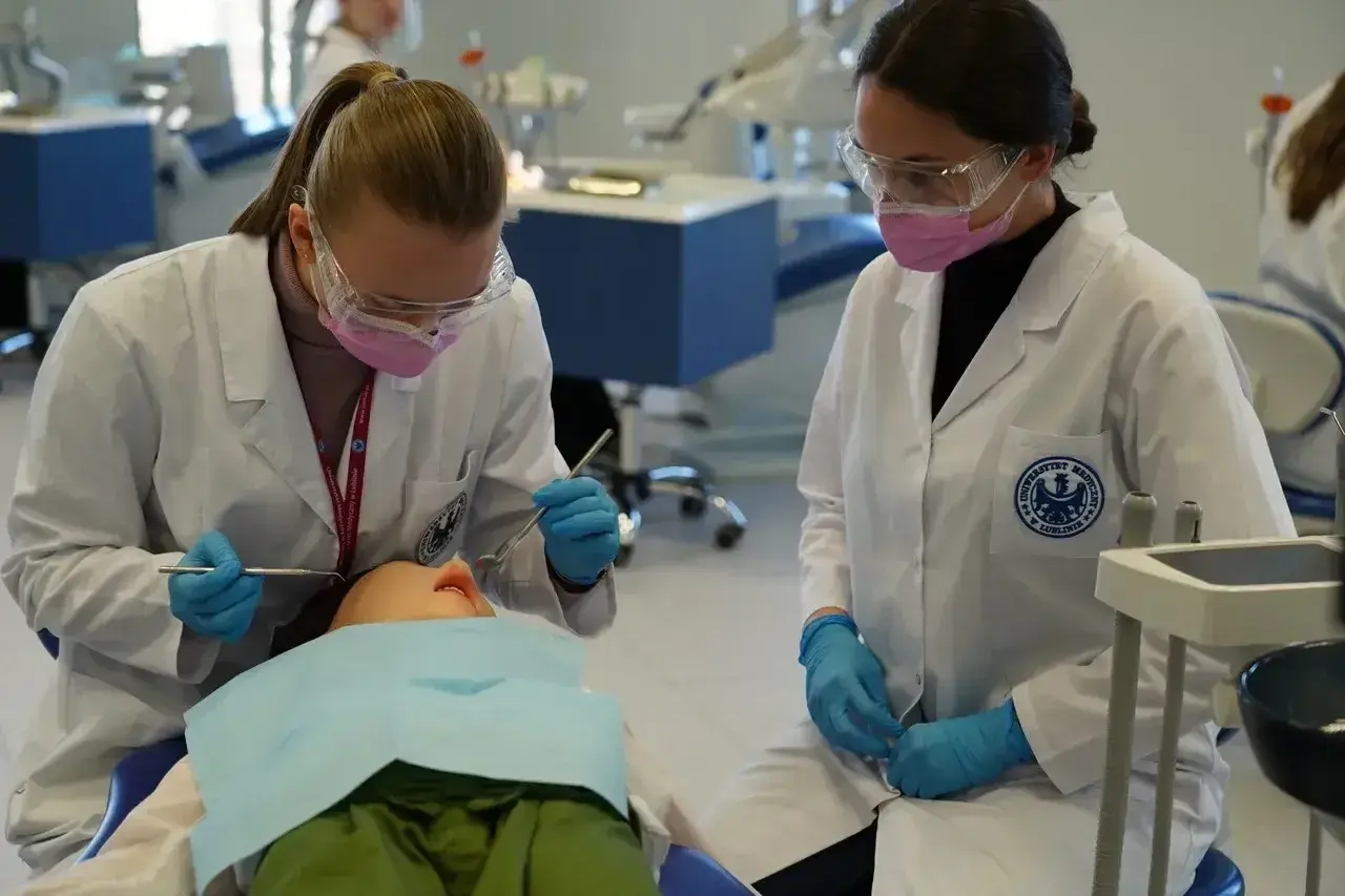 Studentki stomatologii ćwiczą na fantomie. W białych fartuchach, rękawiczkach i okularach ochronnych, z precyzją wykonują zabieg.