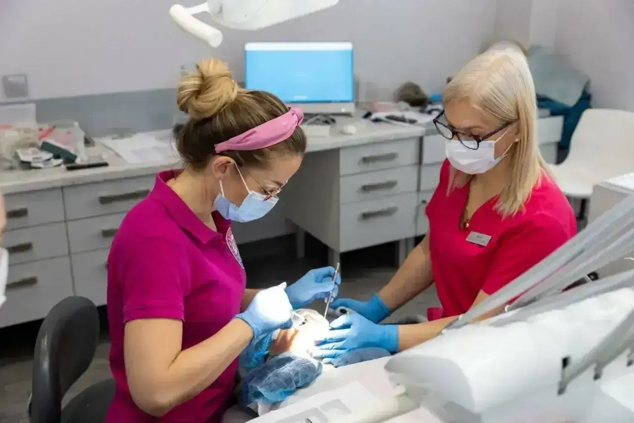 Dwie dentystki w różowych uniformach pracują przy pacjencie. Jedna z nich, z kitką, wykonuje zabieg, druga asystuje. To gabinet, gdzie dba się o zdrowie zębów, a artykaina pomaga w bezbolesnym leczeniu.
