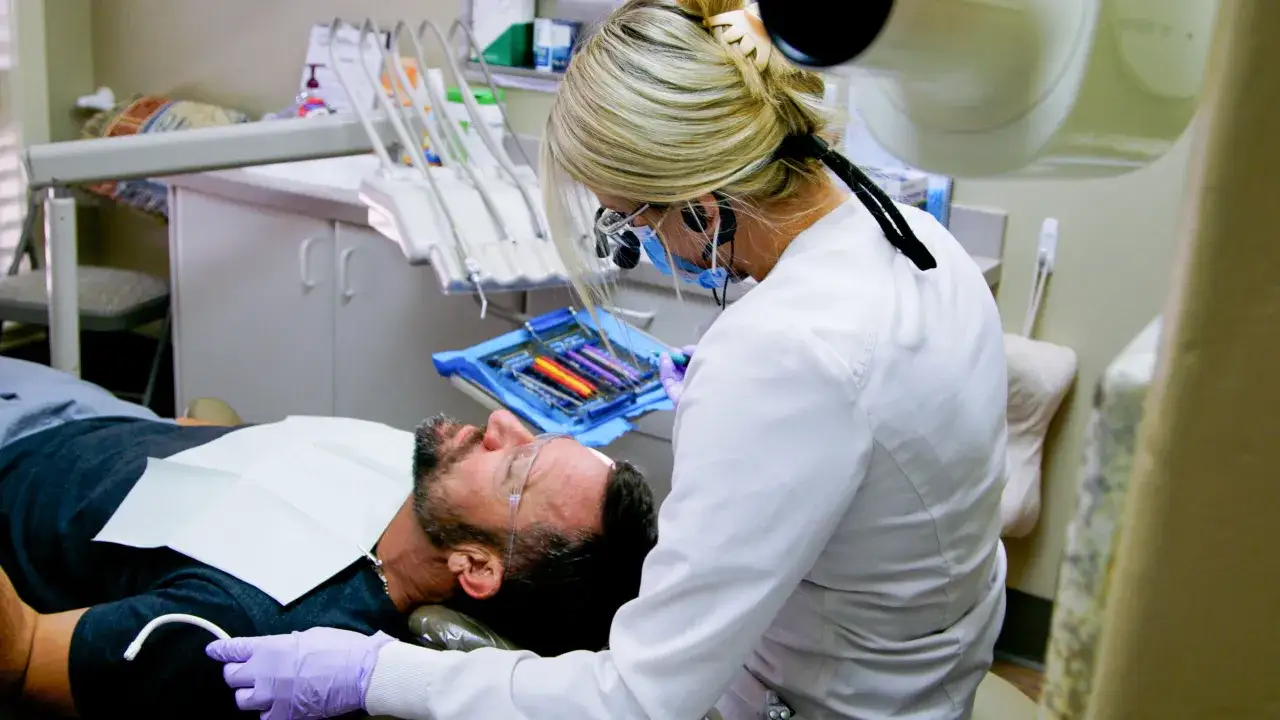 Dentystka w okularach i masce przygotowuje narzędzia. Pacjent leży na fotelu, gotowy na zabieg. Artykaina zapewni komfort.