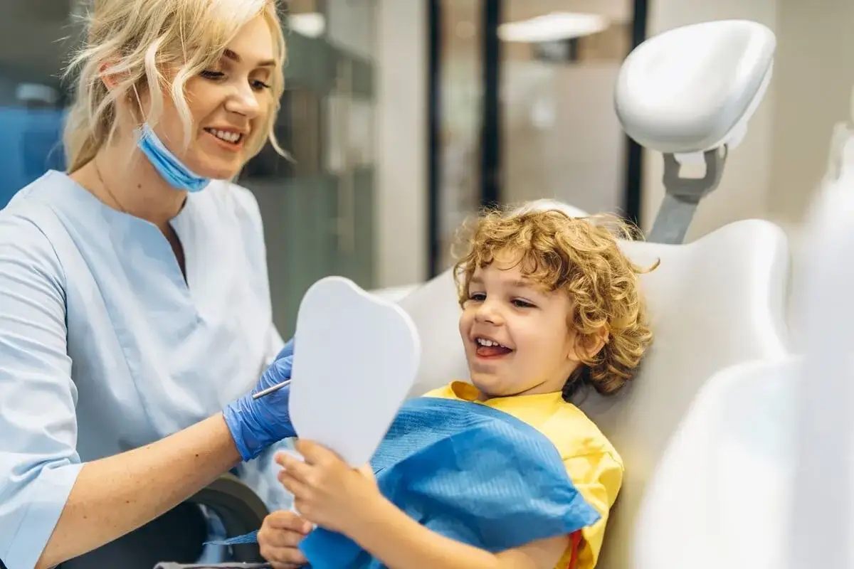 Uśmiechnięty chłopiec z kręconymi włosami ogląda swoje zęby w lusterku, podczas gdy dentystka wyjaśnia mu zasady higieny jamy ustnej. To przykład profesjonalnej pedodoncji.