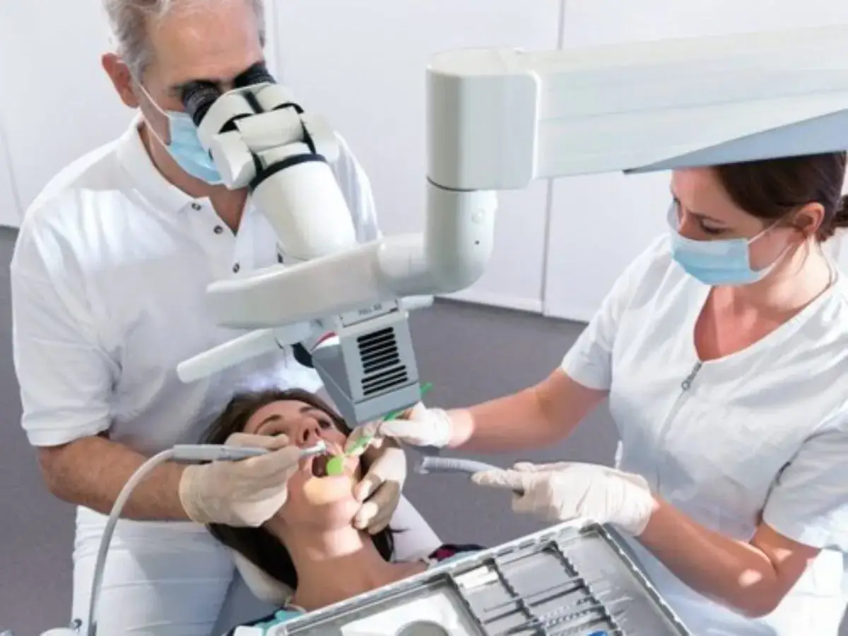 Dentysta wykonuje leczenie kanałowe pod mikroskopem, asystentka przygotowuje narzędzia.