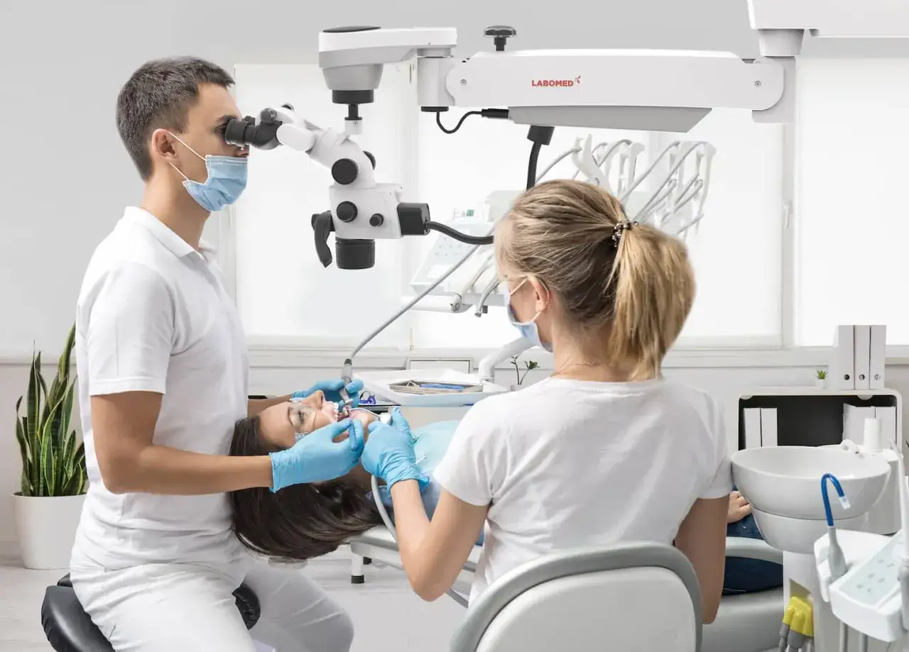 Dentysta wykonuje leczenie kanałowe pod mikroskopem, precyzyjnie pracując nad zębem pacjentki. Asystentka wspiera zabieg.