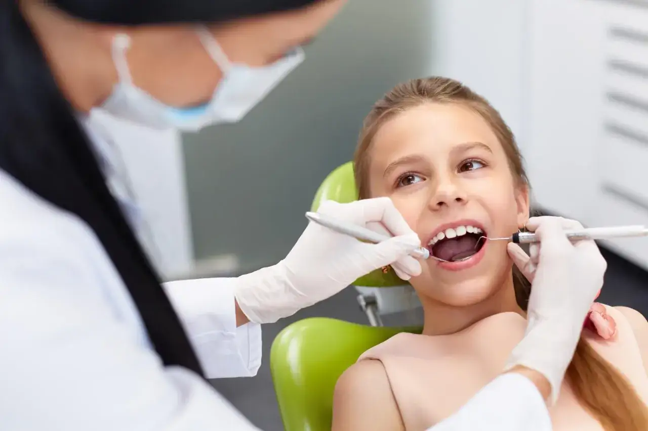 Dziewczynka uśmiecha się podczas wizyty u dentysty. Specjalista pedodoncji bada jej zęby.