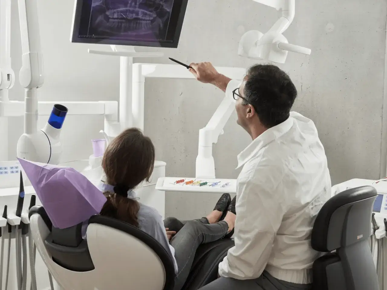 Dentysta omawia z pacjentką plan leczenia, wskazując na RTG zębów. Widoczne narzędzia sugerują przygotowania do ekstrakcji zęba.